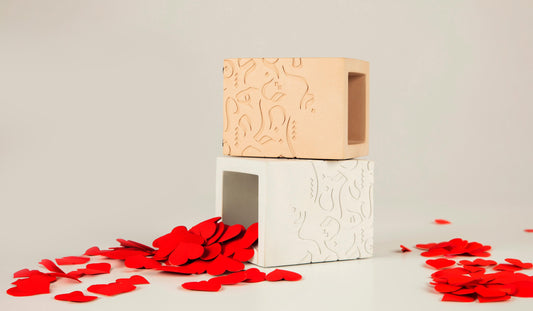 Two textured concrete vessels, one beige and one white, with abstract patterns. Stacked with vibrant red heart-shaped cutouts spilling around, symbolising love and refined luxury.