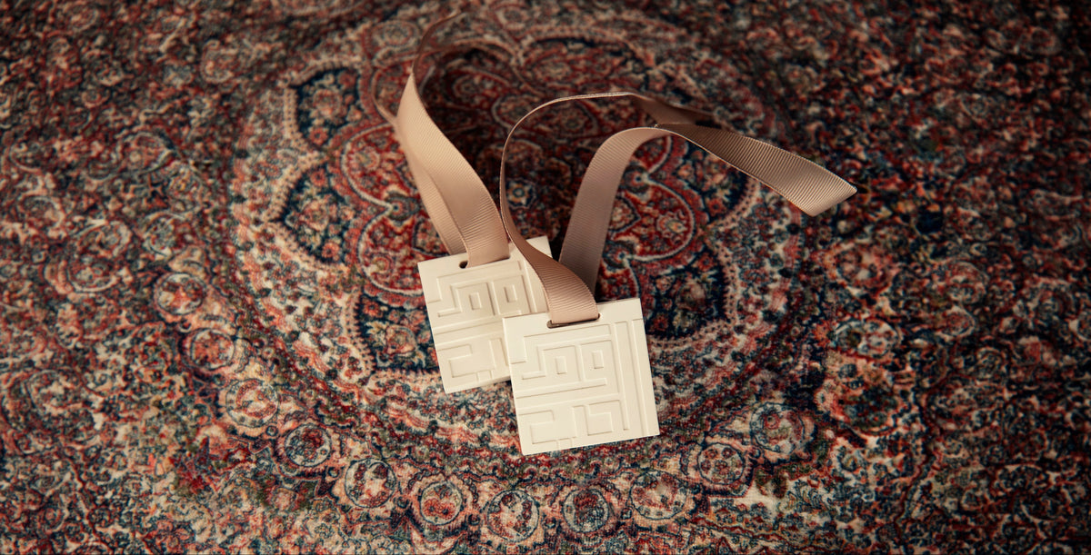 Two square cream-colored ceramic tags with embossed Greek key patterns, attached to tan ribbons, resting on a detailed red and blue patterned rug.