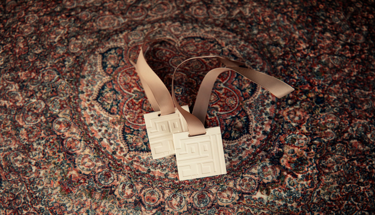 Two square cream-colored ceramic tags with embossed Greek key patterns, attached to tan ribbons, resting on a detailed red and blue patterned rug.